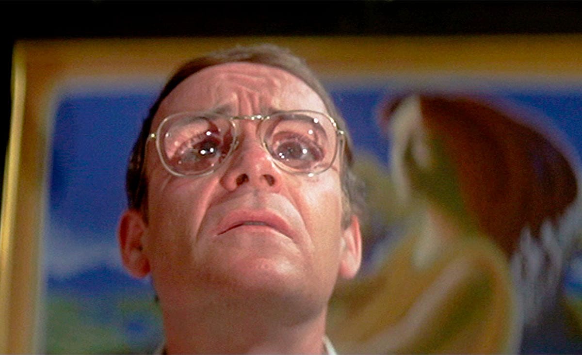Buck Henry as Oliver Farnsworth in The Man Who Fell to Earth (1976), wearing large eyeglasses and looking upward with a worried expression.