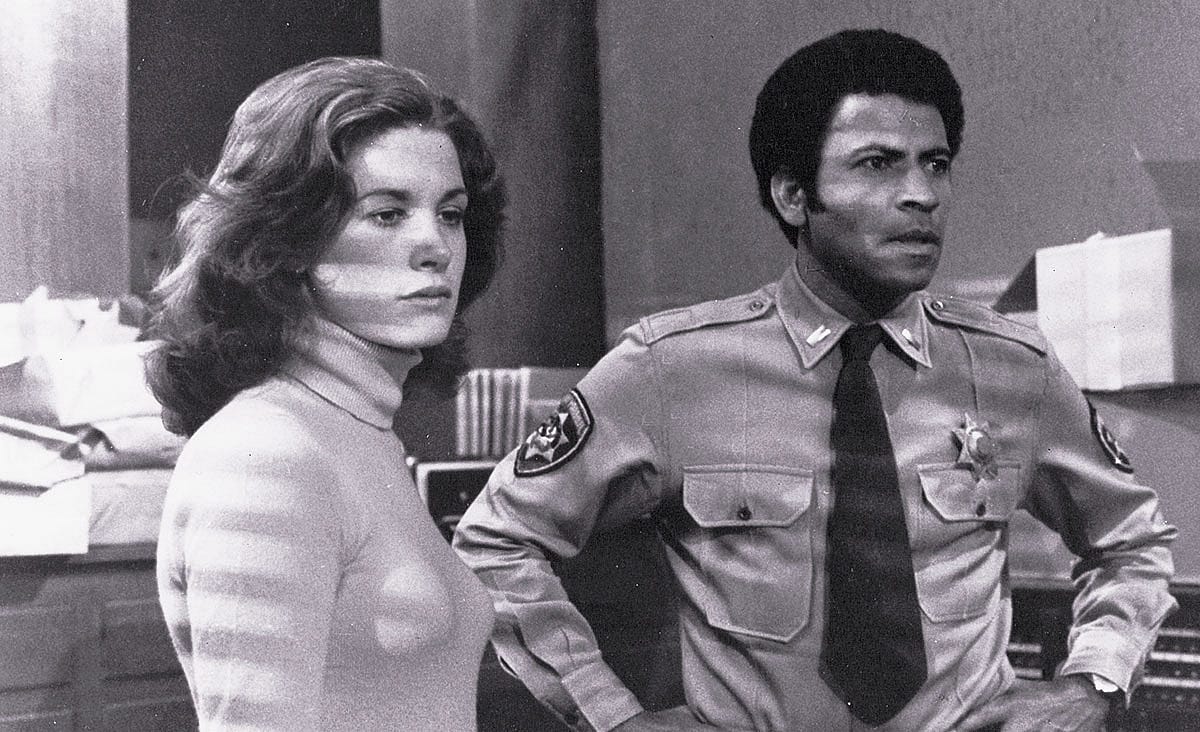 Austin Stoker and Laurie Zimmer in Assault on Precinct 13 (1976), standing together inside a police station, with Stoker wearing a sheriff's uniform and Zimmer dressed in a turtleneck sweater.