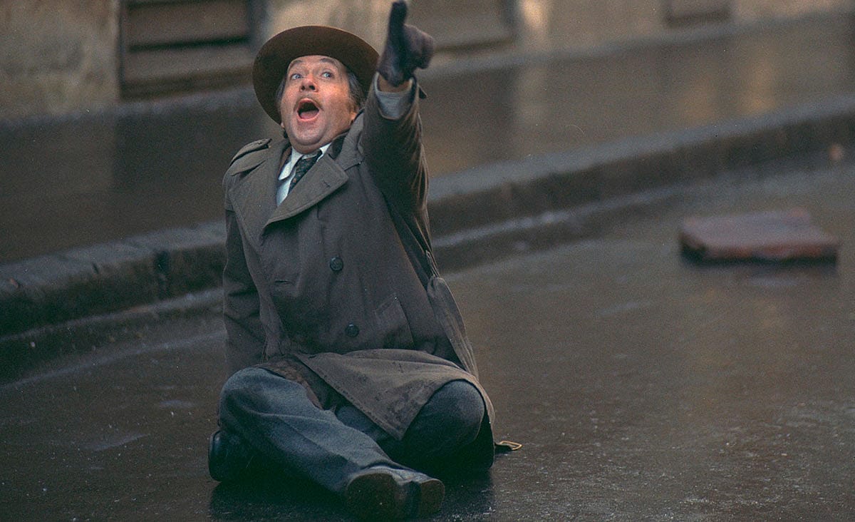 A man in a trench coat and wide-brimmed hat sits on a rain-soaked street, pointing upward with his mouth open, in a dramatic scene from The Fifth Seal (Az ötödik pecsét, 1976).