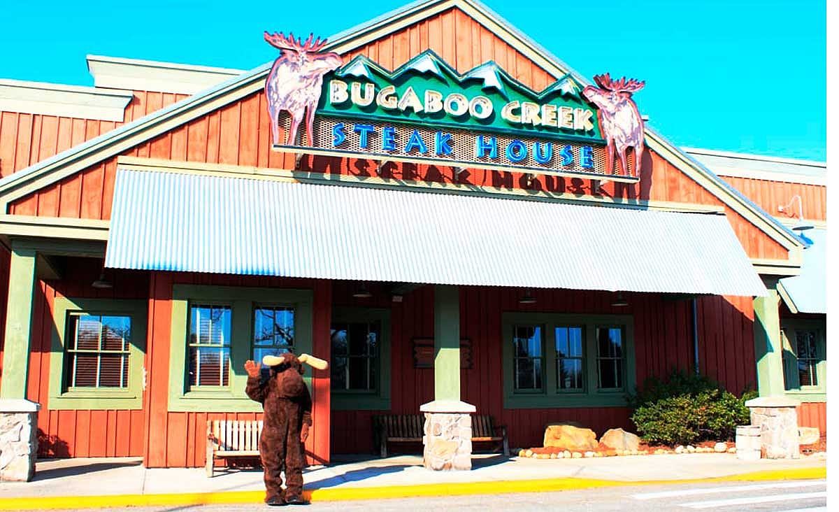 Exterior of a Bugaboo Creek Steak House restaurant with a moose mascot standing outside the entrance beneath the restaurant's mountain-themed sign.