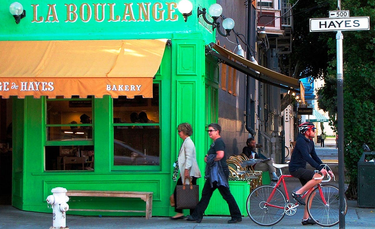 Exterior of a La Boulange bakery on a city street corner with pedestrians walking by and a cyclist stopped outside.
