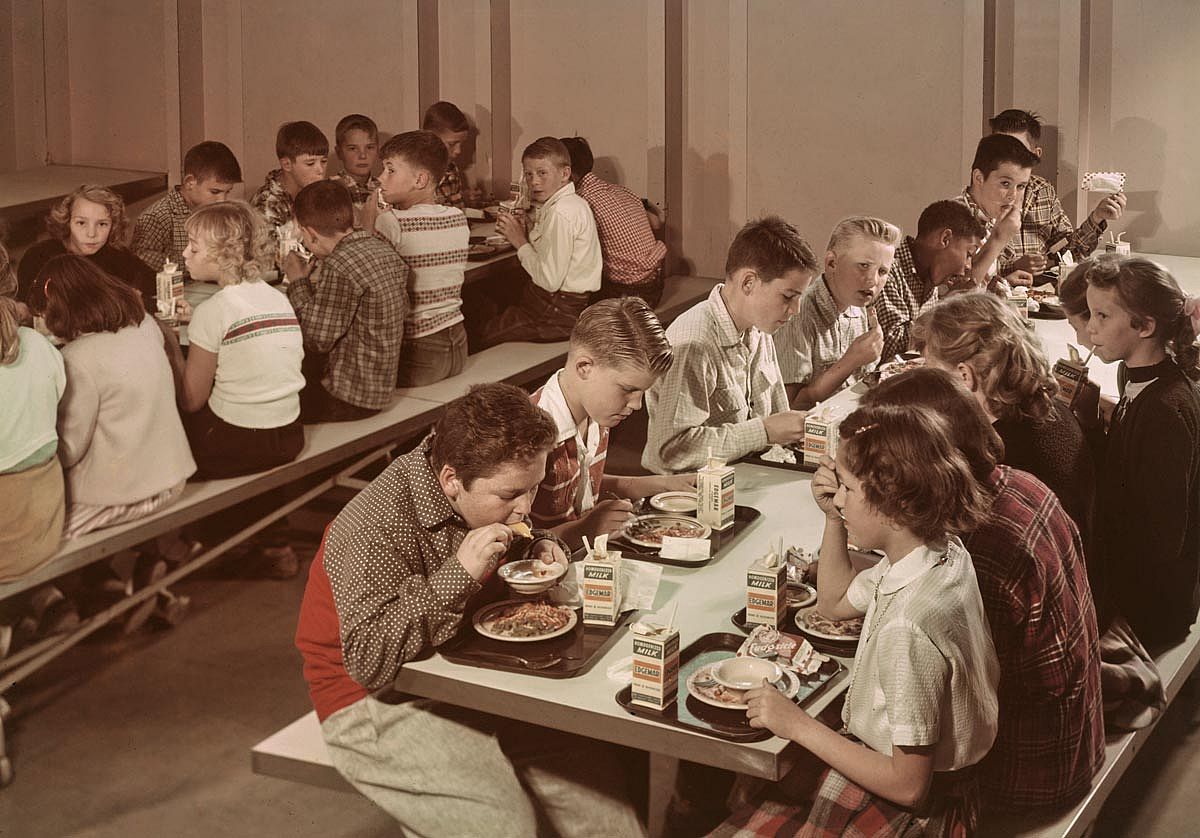 Vintage color photo of schoolchildren seated at long cafeteria tables eating lunch from trays, circa 1965.
