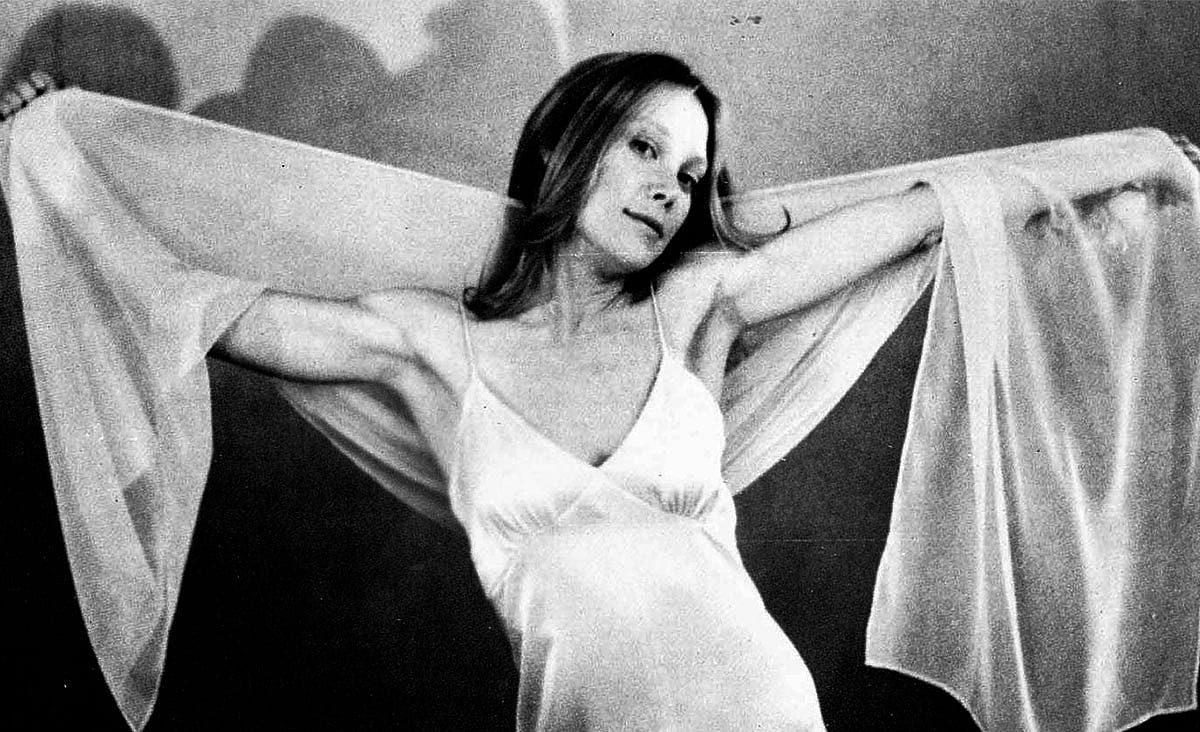 Sissy Spacek as Carrie White in Carrie (1976), wearing a flowing white nightgown with sheer sleeves and posing with her arms outstretched in a black-and-white production still.