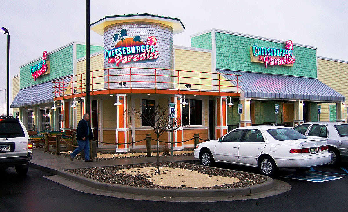 Exterior of a Cheeseburger in Paradise restaurant with neon signs, parked cars, and a pedestrian walking toward the entrance.