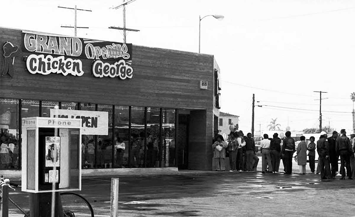 Crowd gathered outside a Chicken George restaurant during its grand opening, with a payphone booth in the foreground.