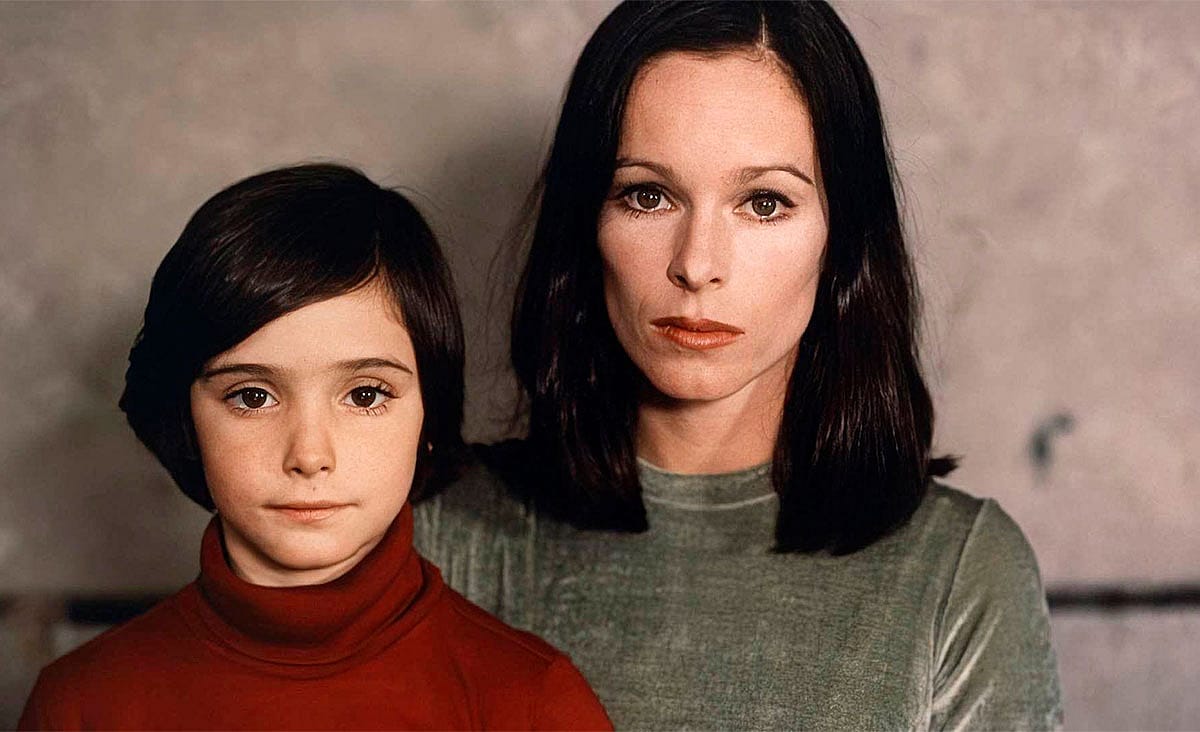 Geraldine Chaplin and Ana Torrent in Cría cuervos (1976), posing side by side with solemn expressions against a plain indoor background.