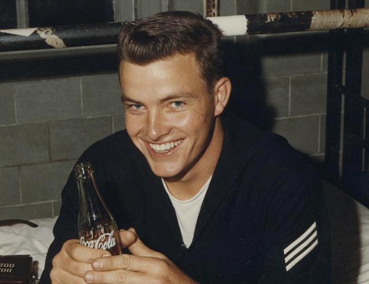A colorized photo of a young man in the Navy smiling for the camera while holding a bottle of Coca-Cola.