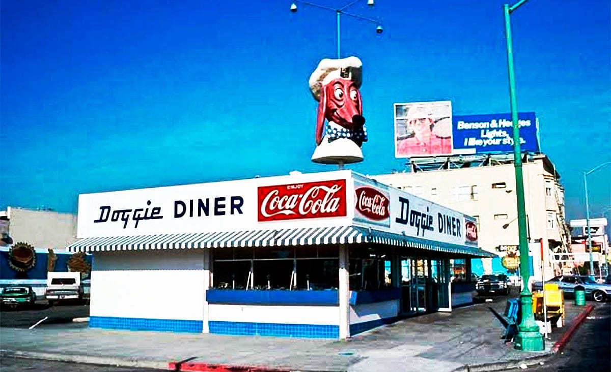 Exterior of the Doggie Diner with a giant dachshund chef mascot sign on the roof and Coca-Cola branding on the building.