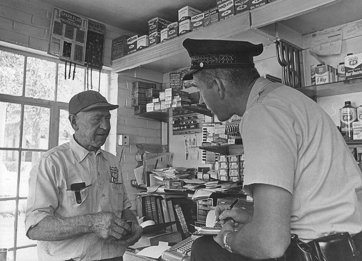Black-and-white 1965 photo of a Phillips 66 gas station owner talking with a police officer inside a cluttered station office, surrounded by shelves of oil filters and car parts.