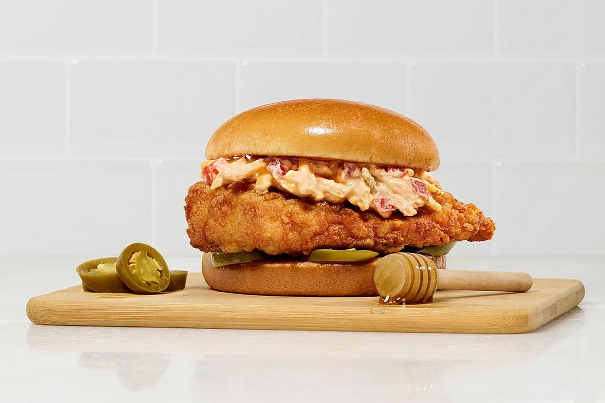 A photo of the honey pepper pimento sandwich with spicy filet from Chick-fil-A sitting on a cutting board with some jalapeños and a honey dipper.