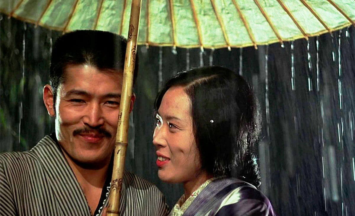 Tatsuya Fuji and Eiko Matsuda in In the Realm of the Senses (Ai no Corrida, 1976), standing together beneath a traditional paper umbrella in the rain.
