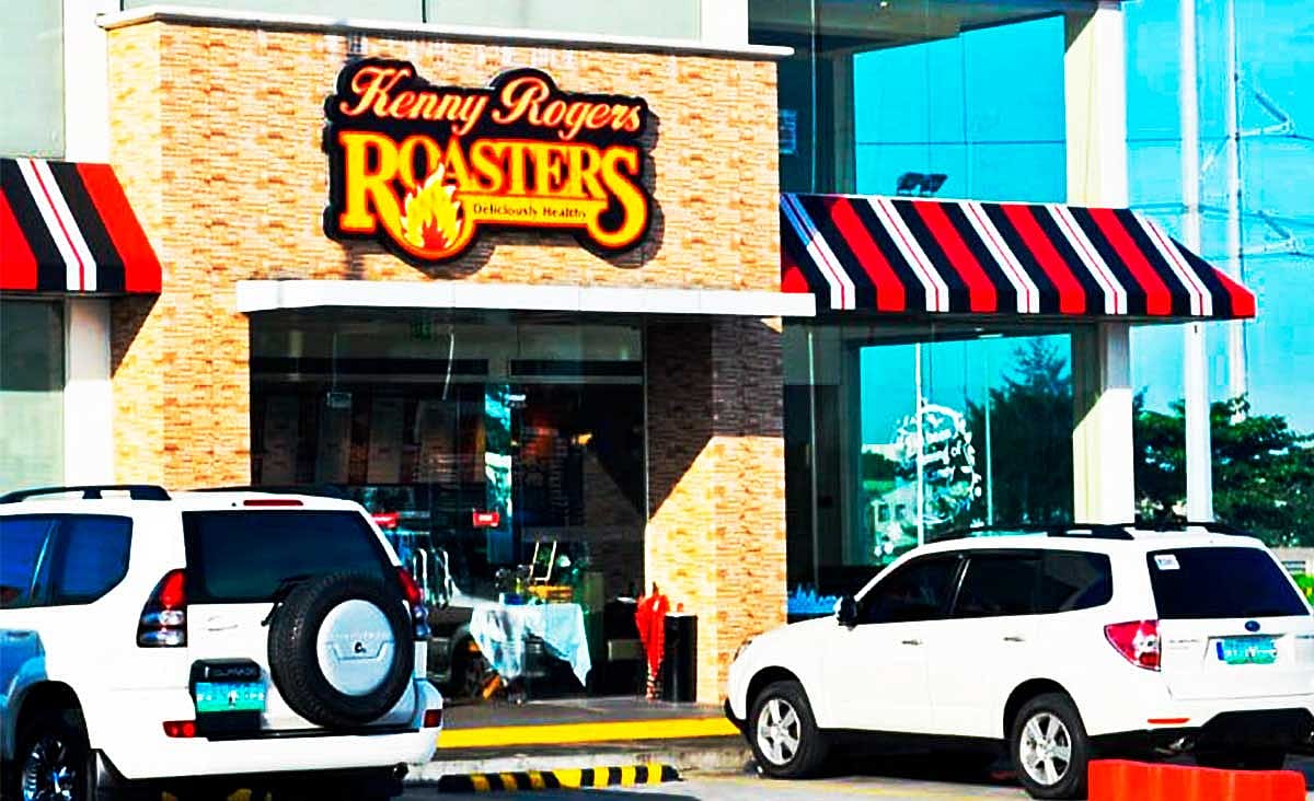 Exterior of a Kenny Rogers Roasters restaurant with striped awnings, parked SUVs, and the restaurant's illuminated sign above the entrance.