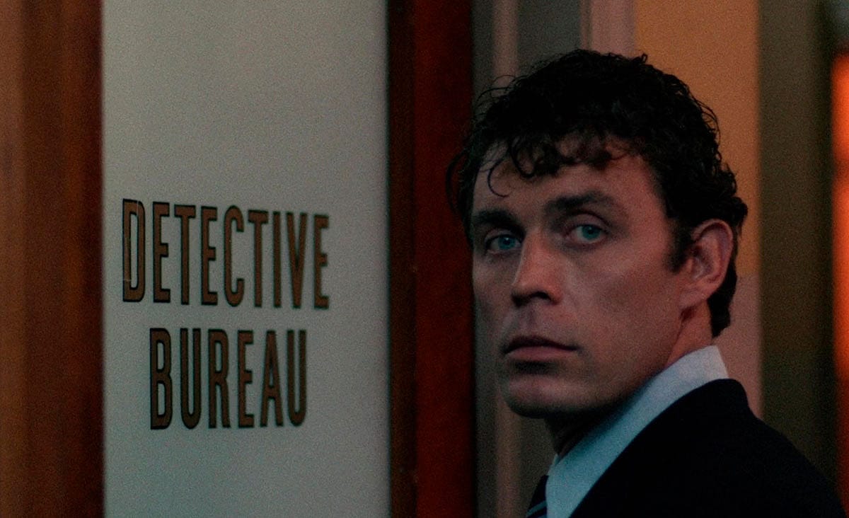 A man in a dark suit looks over his shoulder beside a door labeled “Detective Bureau” in a scene from Alice, Sweet Alice (1976).