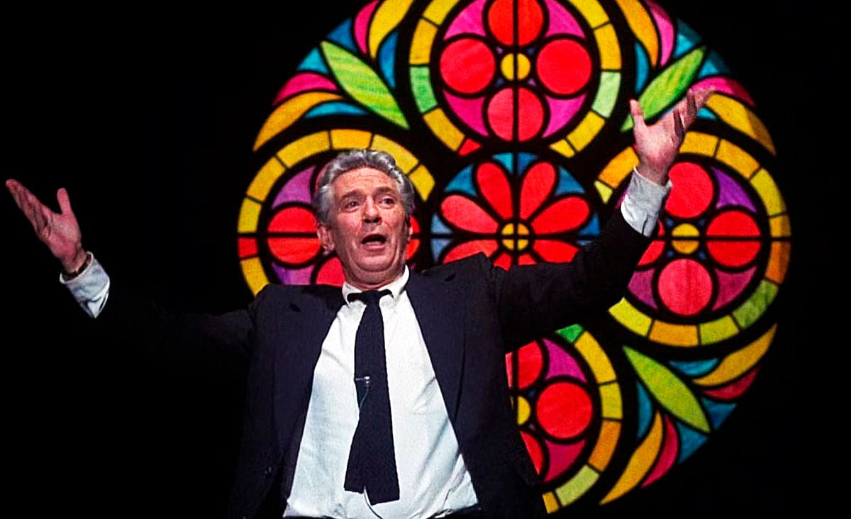 Peter Finch as Howard Beale in Network (1976), standing with his arms outstretched in front of a colorful stained-glass window while delivering an impassioned speech.