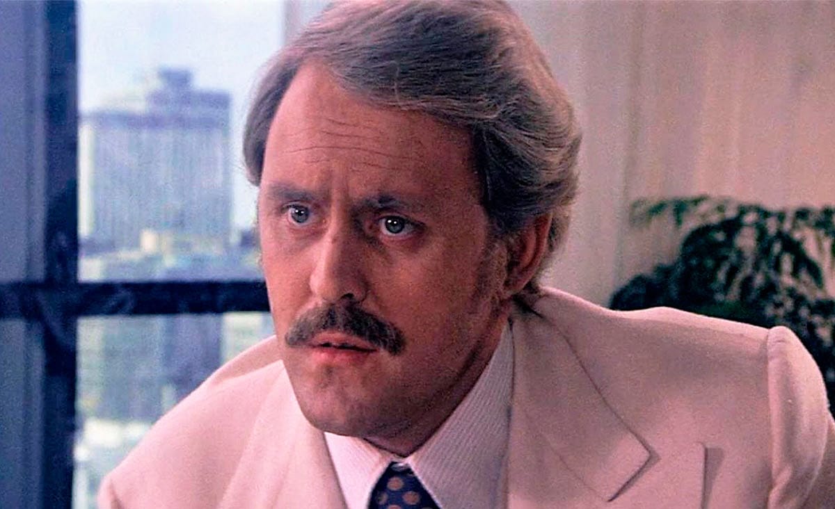American actor Cliff Robertson in Obsession (1976), wearing a light-colored suit and patterned tie while looking ahead inside a modern office with city buildings visible through the window.