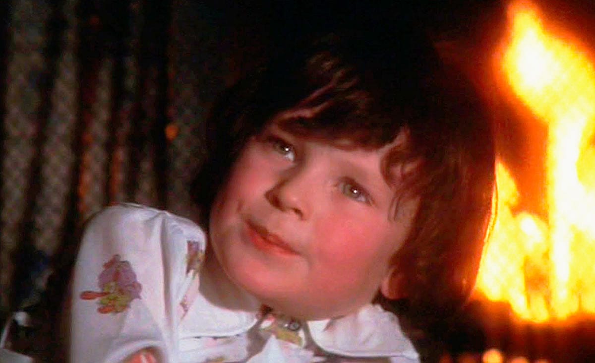Harvey Spencer Stephens as Damien Thorn in The Omen (1976), wearing patterned pajamas and looking upward while sitting near a glowing fireplace.