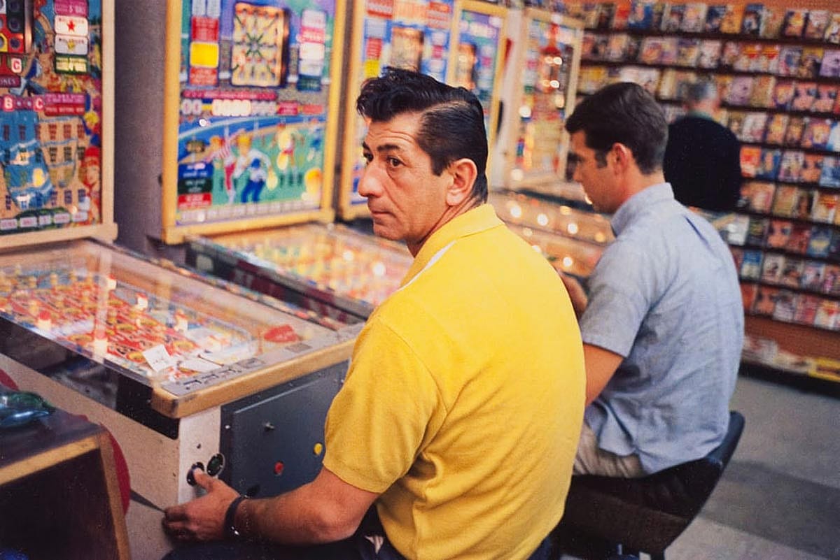 Two men sit at Pinball machines in a beautiful color photo