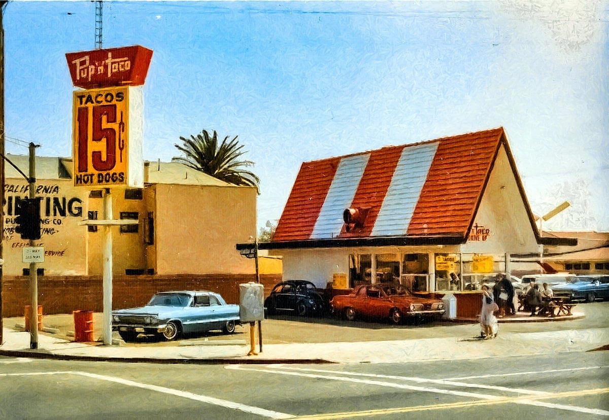Vintage Pup 'N' Taco restaurant with a roadside sign advertising 15-cent tacos and hot dogs, surrounded by classic cars.