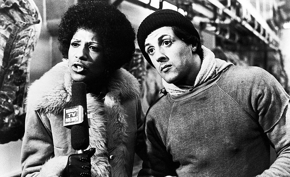 Sylvester Stallone and Jayne Kennedy in Rocky (1976), standing together at a meatpacking facility as Kennedy holds a television news microphone.
