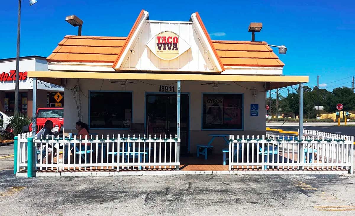 Exterior of a Taco Viva fast-food restaurant with outdoor patio seating enclosed by a white picket fence.