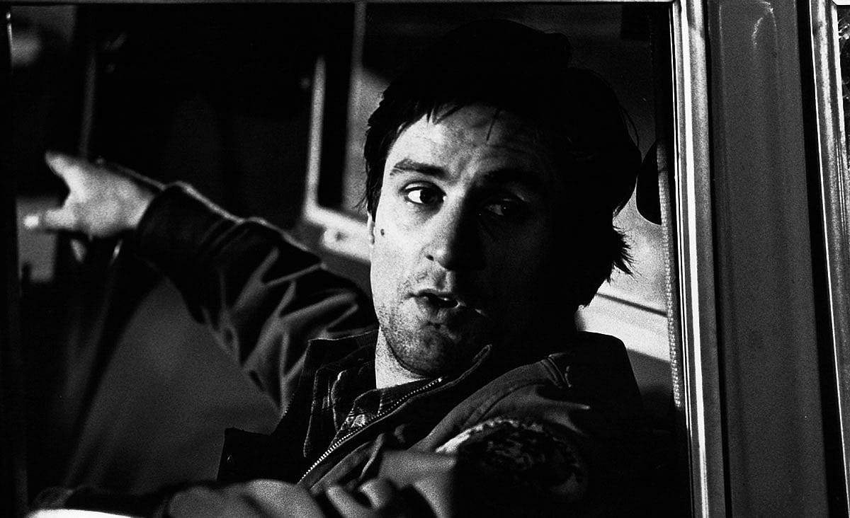 Robert De Niro as Travis Bickle in Taxi Driver (1976), seated inside a taxi with one arm resting on the open window, glancing over his shoulder in a black-and-white image.