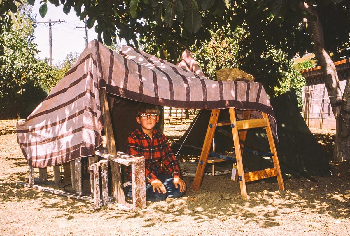 A young boy with glasses sitting under a fort he made in his backyard.