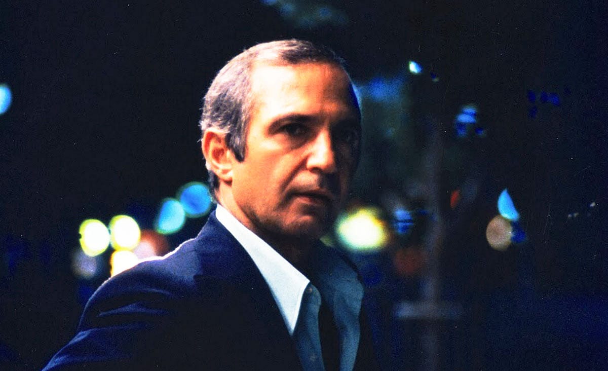 Ben Gazzara in The Killing of a Chinese Bookie (1976), wearing a dark suit and looking over his shoulder on a city street at night with blurred lights in the background.