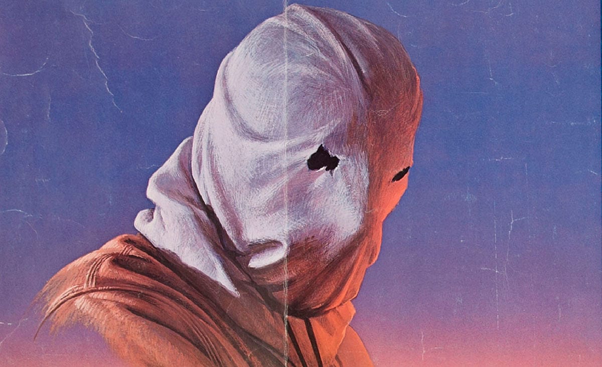 The masked killer from The Town That Dreaded Sundown (1976), wearing a white burlap hood with eye holes and a brown jacket against a blue-purple background.