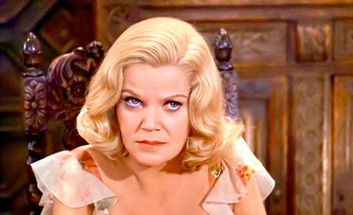 American actress Eileen Brennan as Tess Skeffington in Murder by Death (1976), seated in an ornate wooden chair with a serious expression inside a lavish mansion.