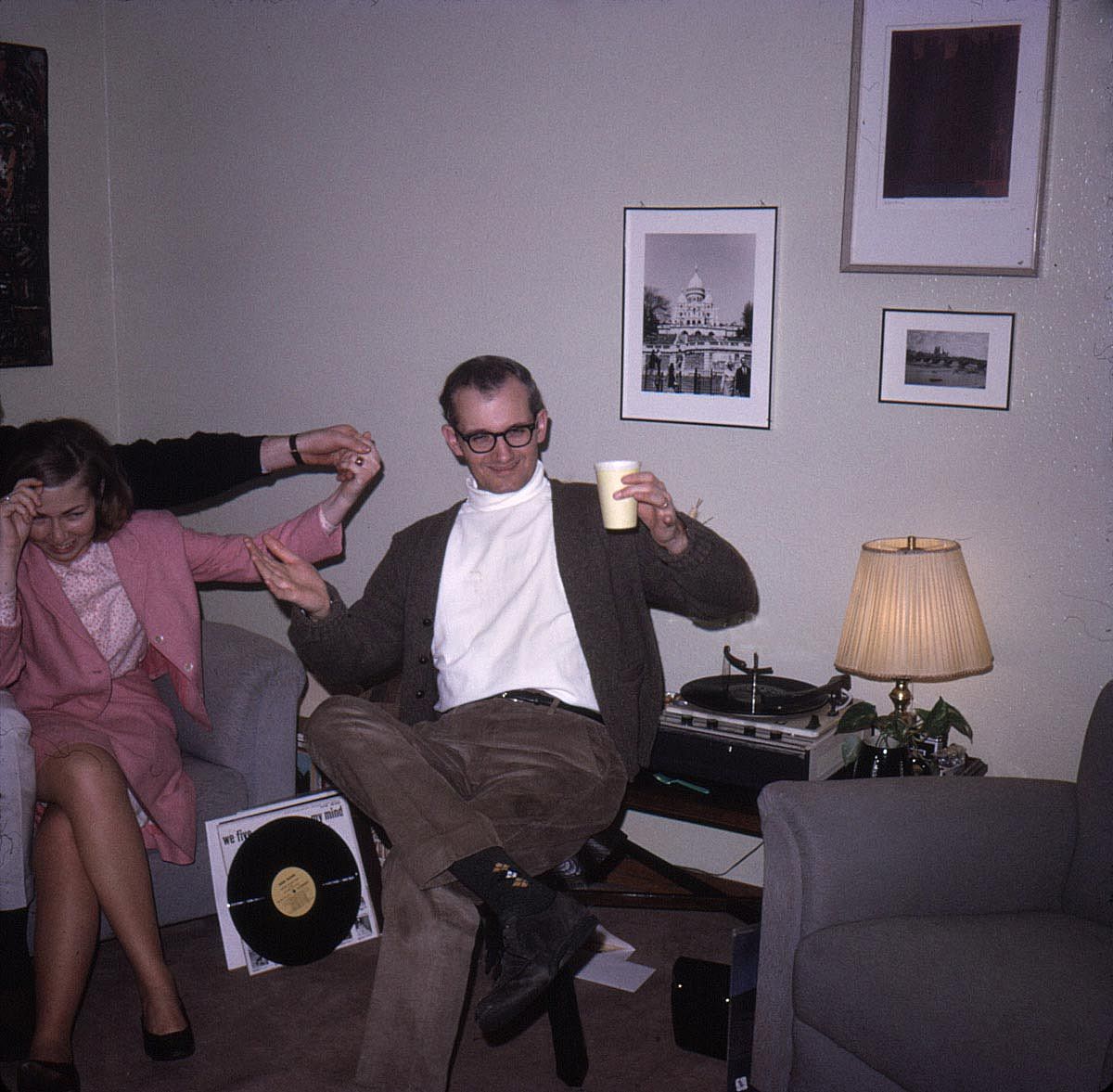 Color photo of a man holding a paper cup and gesturing to the camera.