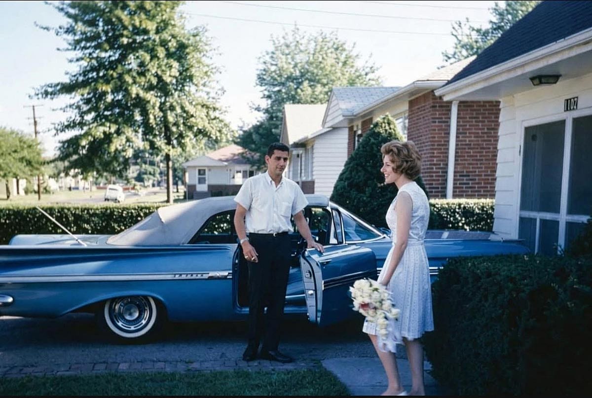 A young woman with a bouquet in her hand and her husband stands nearby holding the passenger side door open for her.