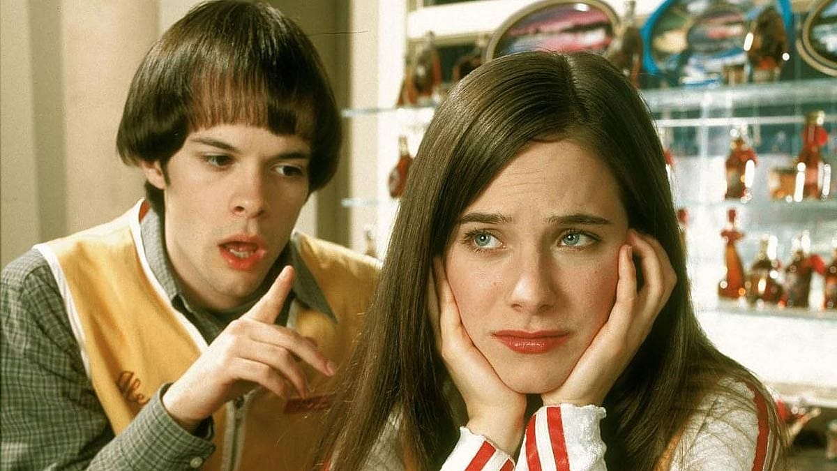 A young woman rests her face in her hands and looks upward with a weary expression while a diner employee gestures and speaks behind her.