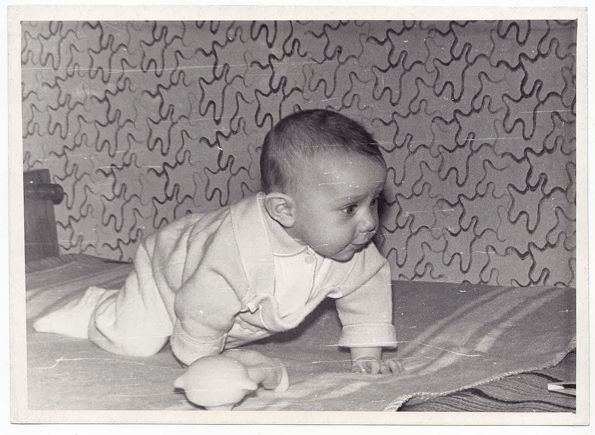 A vintage photograph depicting a baby lying on a blanket. The photo conveys the warm atmosphere of yesteryear. 