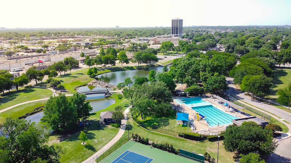 Tennis court and swimming pool sport complex in recreation park with small pond and downtown Richardson, Texas in background. Mature green trees along the trails in summertime