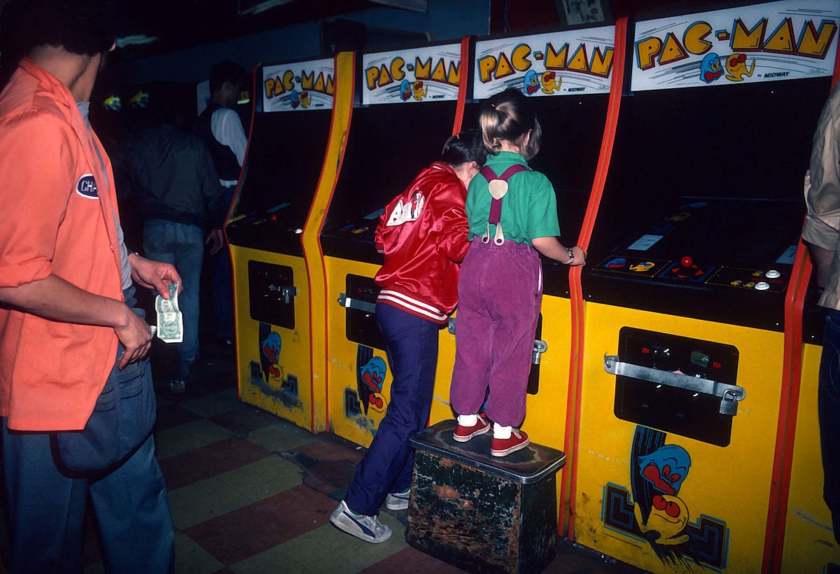 Two young girls play Pac-Man at a row of arcade cabinets in a Times Square video arcade, one standing on a step stool to reach the controls, June 1982.