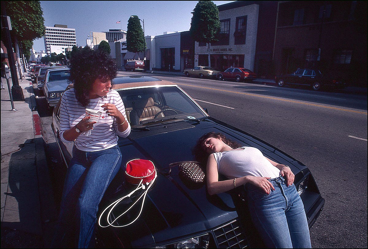 Two teenage girls sit and lie on the hood of a parked car on a Beverly Hills street, one eating from a red thermos cup, 1982.