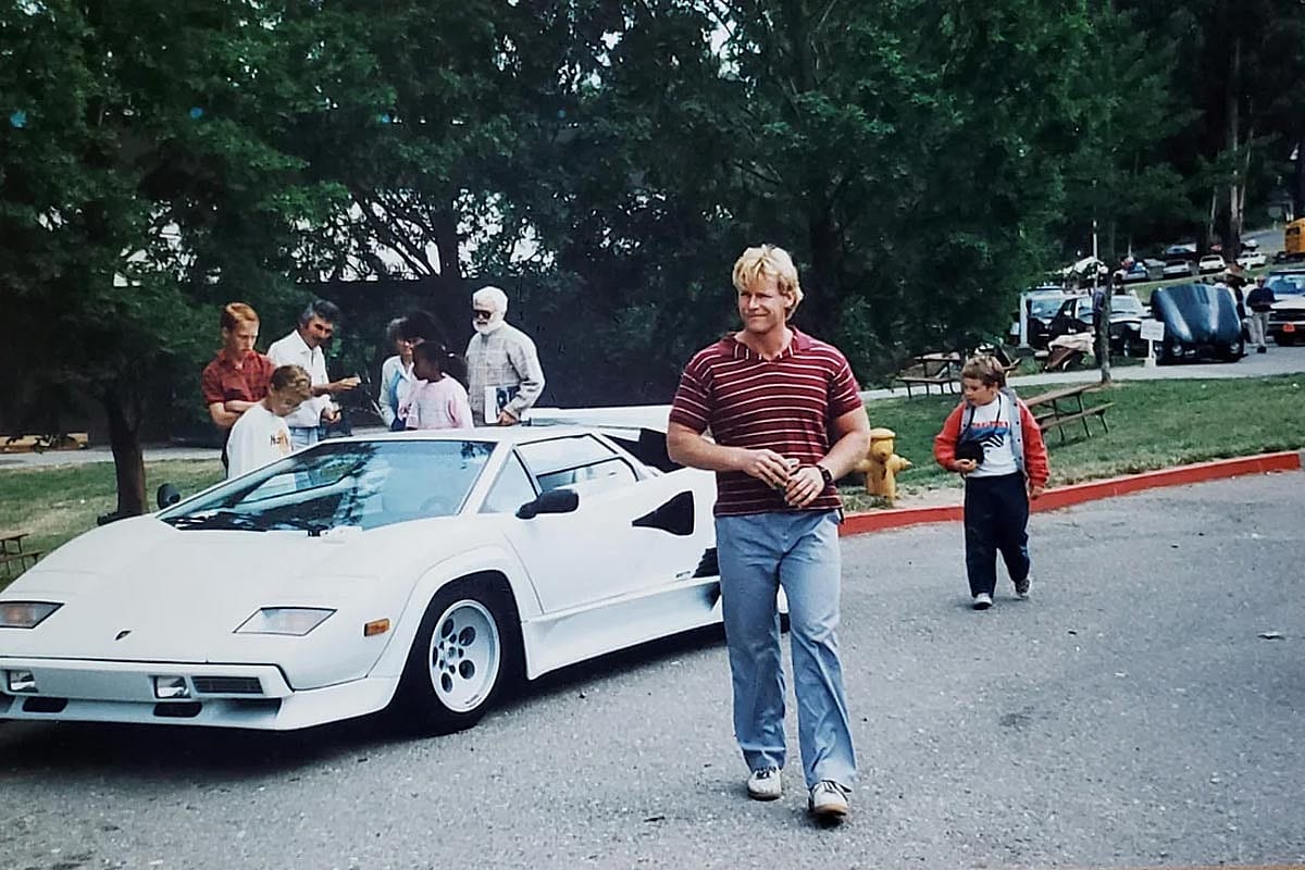 White Lamborghini Countach with scissor doors and pop-up headlights parked on a street, with a man in a striped polo shirt walking away and a small crowd gathered nearby.