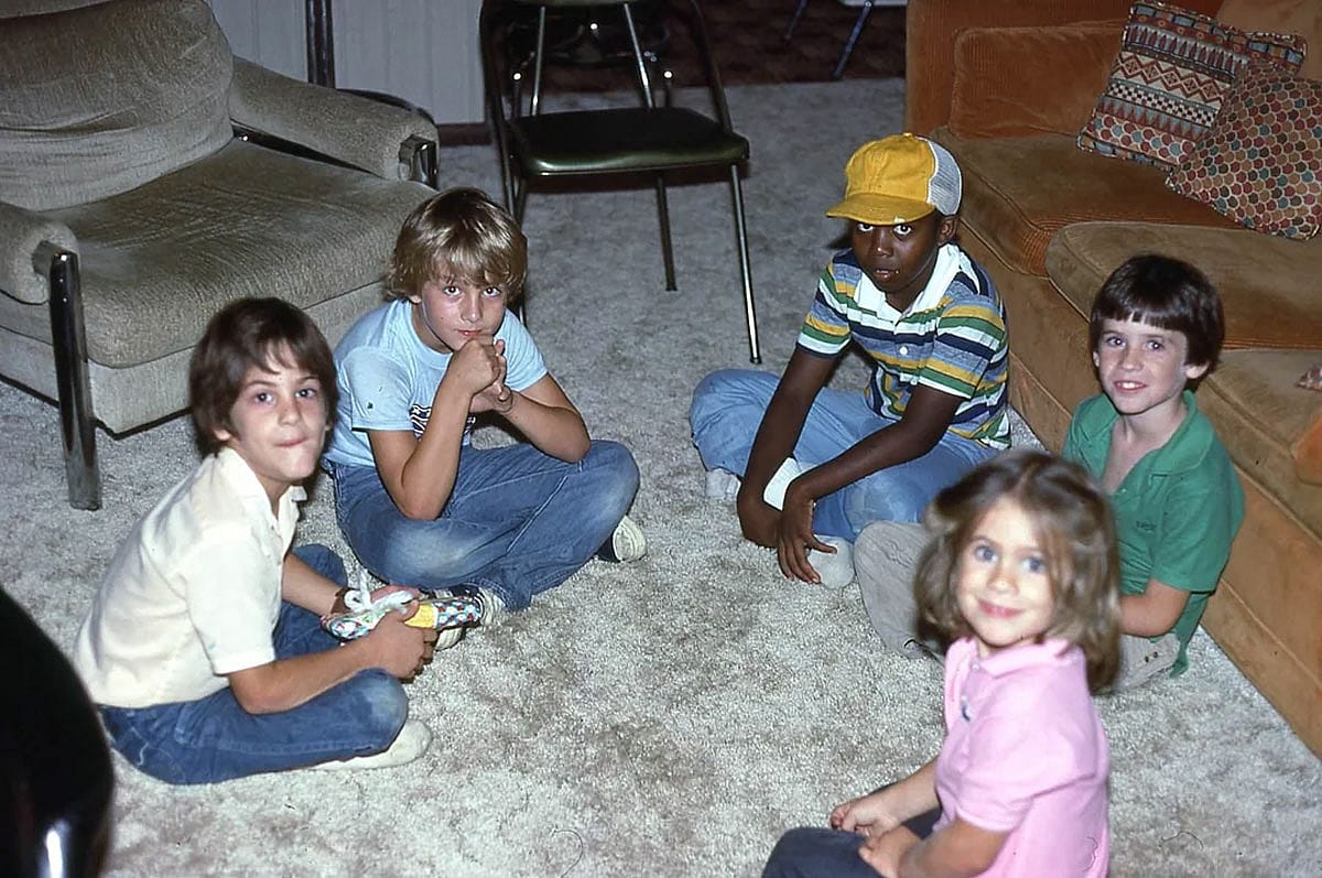 Five kids sit together on a shag carpet in a wood-paneled 1980s living room, 1982.