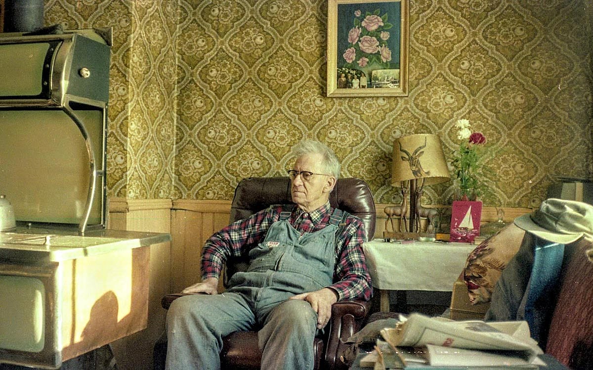 Elderly man in denim overalls and a plaid shirt sits in a brown recliner beside an antique wood-burning stove, in a room with patterned gold wallpaper.