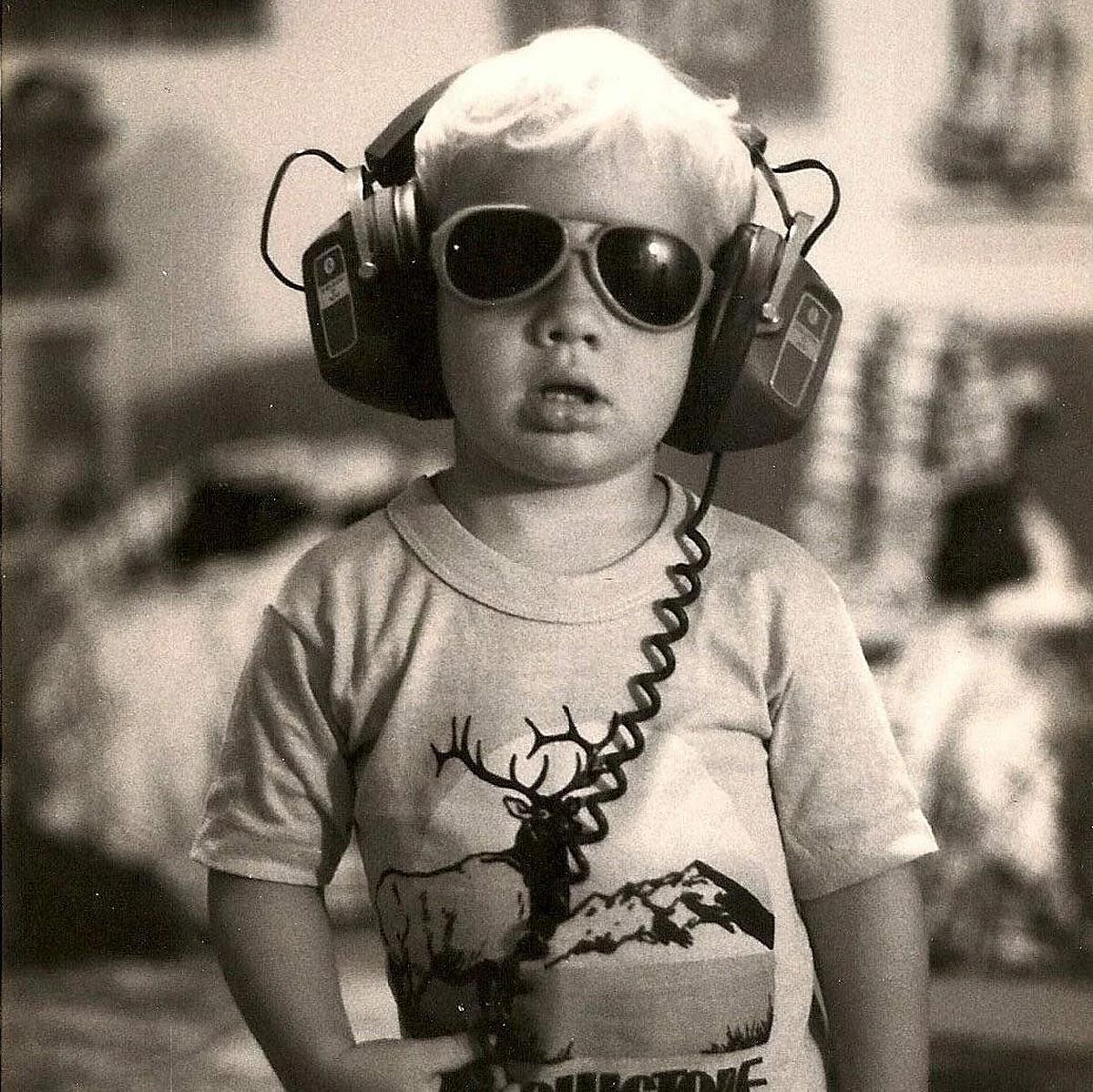 Black-and-white photo of a young child wearing large aviator sunglasses and oversized over-ear headphones, in a T-shirt printed with an elk and mountain scene.