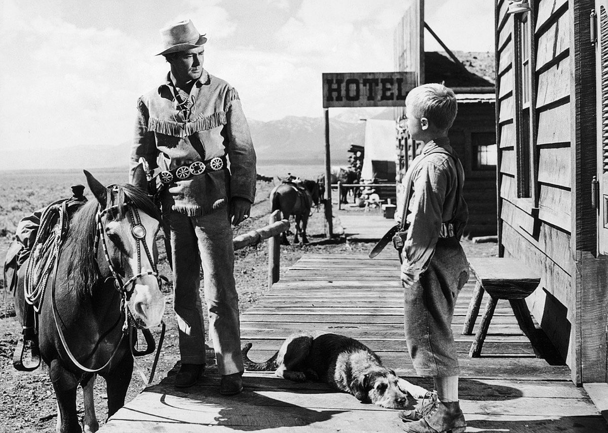 A man in a western hat and fringed jacket talks to a little boy with a dog.