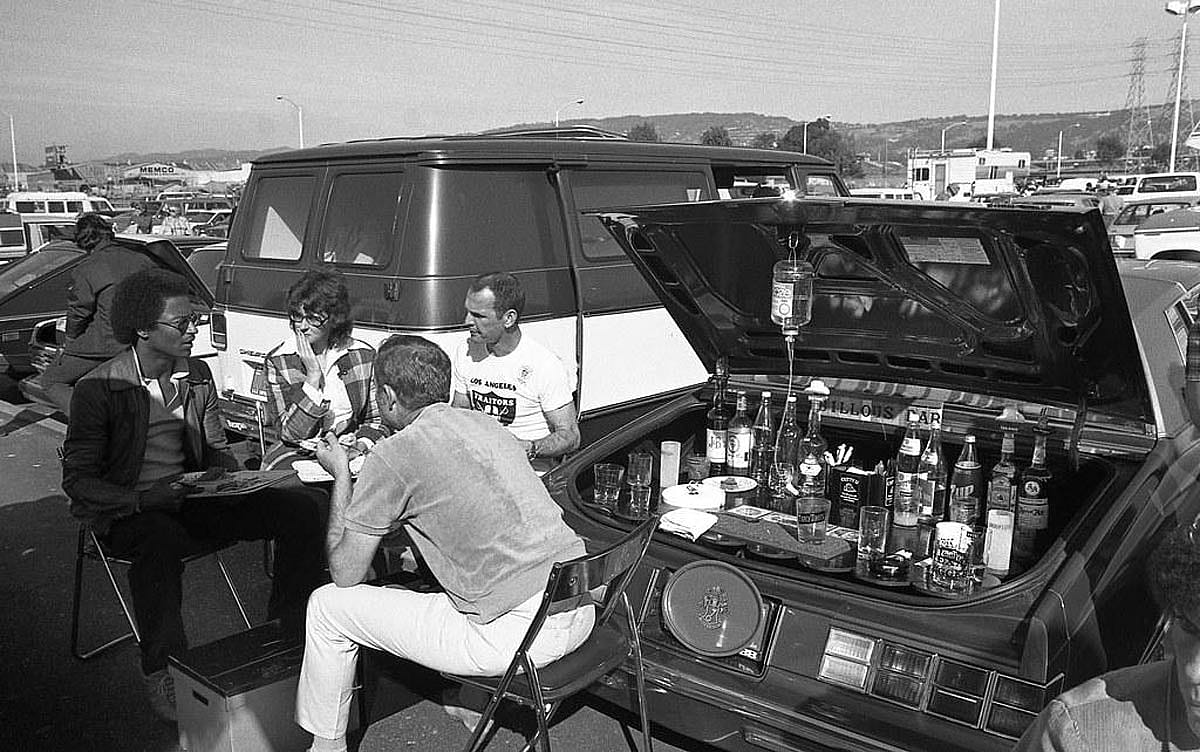 Four people sit in folding chairs around the open trunk of a car set up as a bar with liquor bottles and glasses, parked in a stadium lot; one man's T-shirt reads "Los Angeles Traitors."