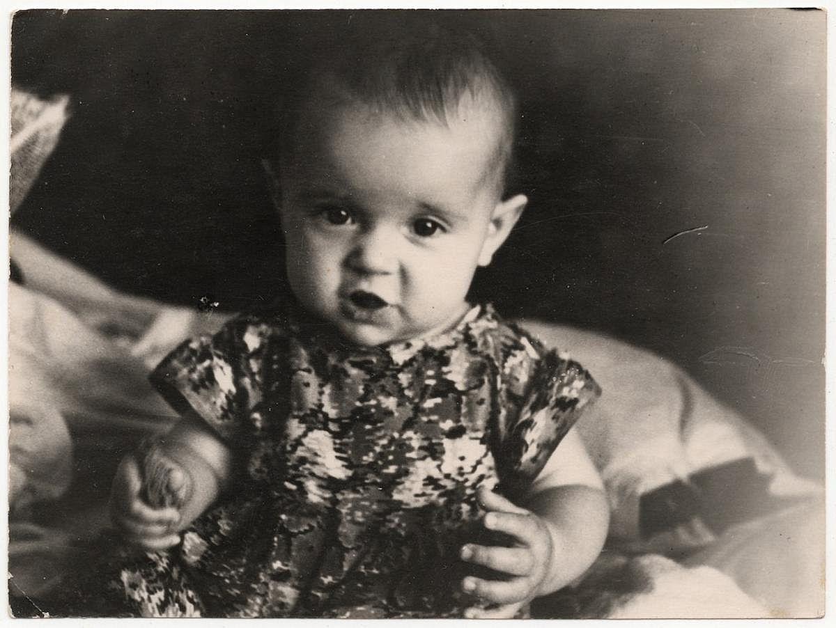 A vintage photograph of a sweet one-year-old baby sitting on her crib indoors.