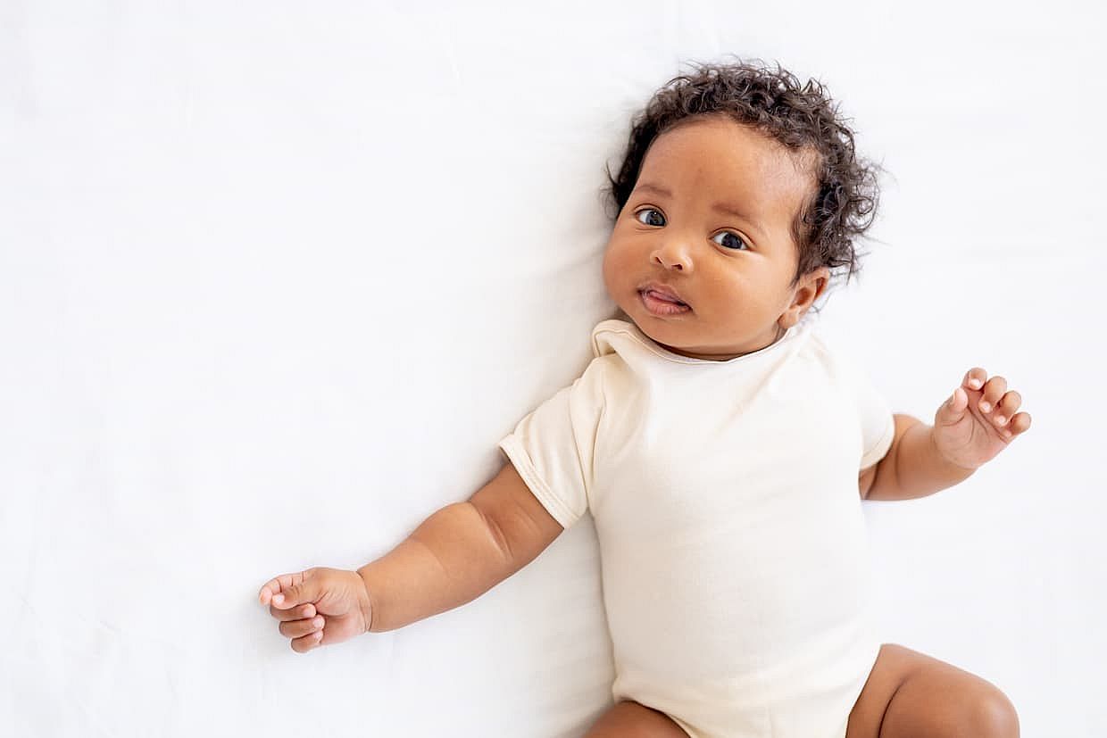 close-up portrait of a small African American baby girl in a white bodysuit on a cotton bed at home, funny six-month-old smiling joyful black newborn baby shows tongue.