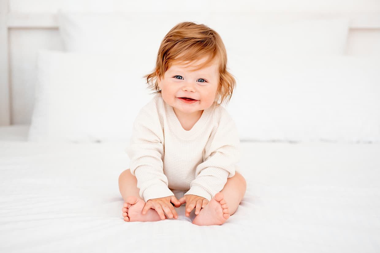 A baby with red hair and blue eyes in a white bodysuit is smiling happily on the bed at home, a healthy happy cute newborn baby boy.