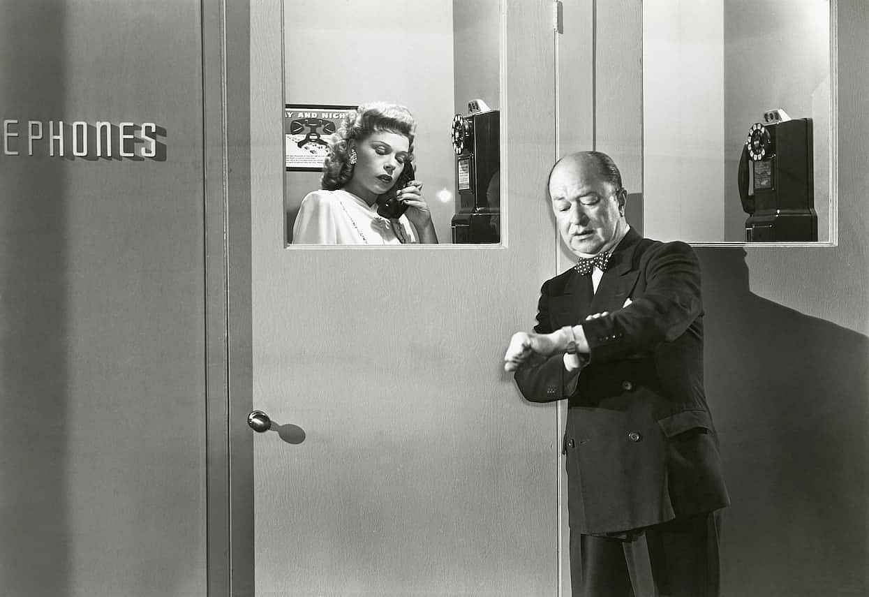 Black and white photo of a woman on a telephone and a man checking his watch while waiting his turn
