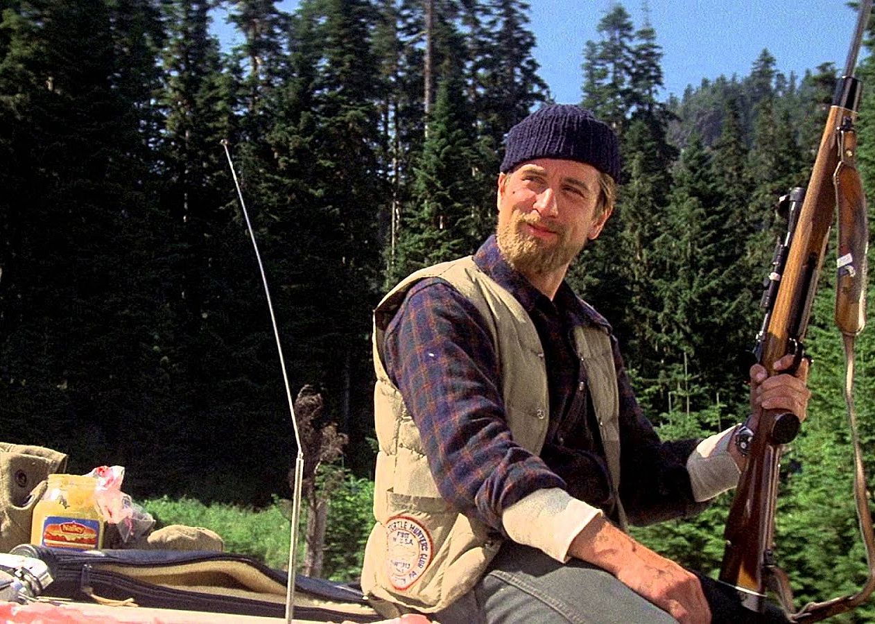 Robert De Niro in a plaid shirt, vest and beanie sitting on the hood of a car holding a rifle.