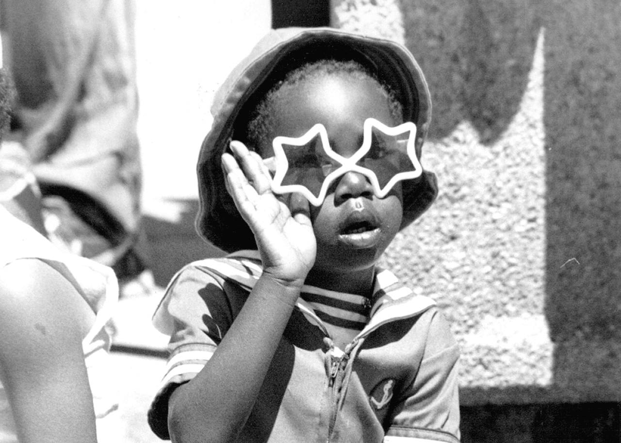 A child wearing star sunglasses waves at a passing parade.