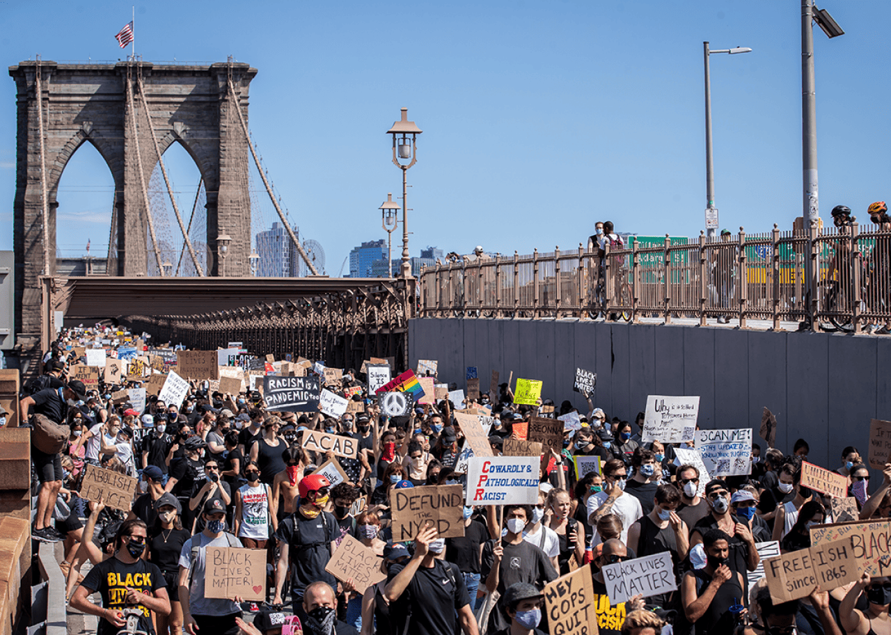 Thousands of protesters walk in a peaceful protest across the Brooklyn Bridge holding signs Brooklyn Bridge Arch in the background.