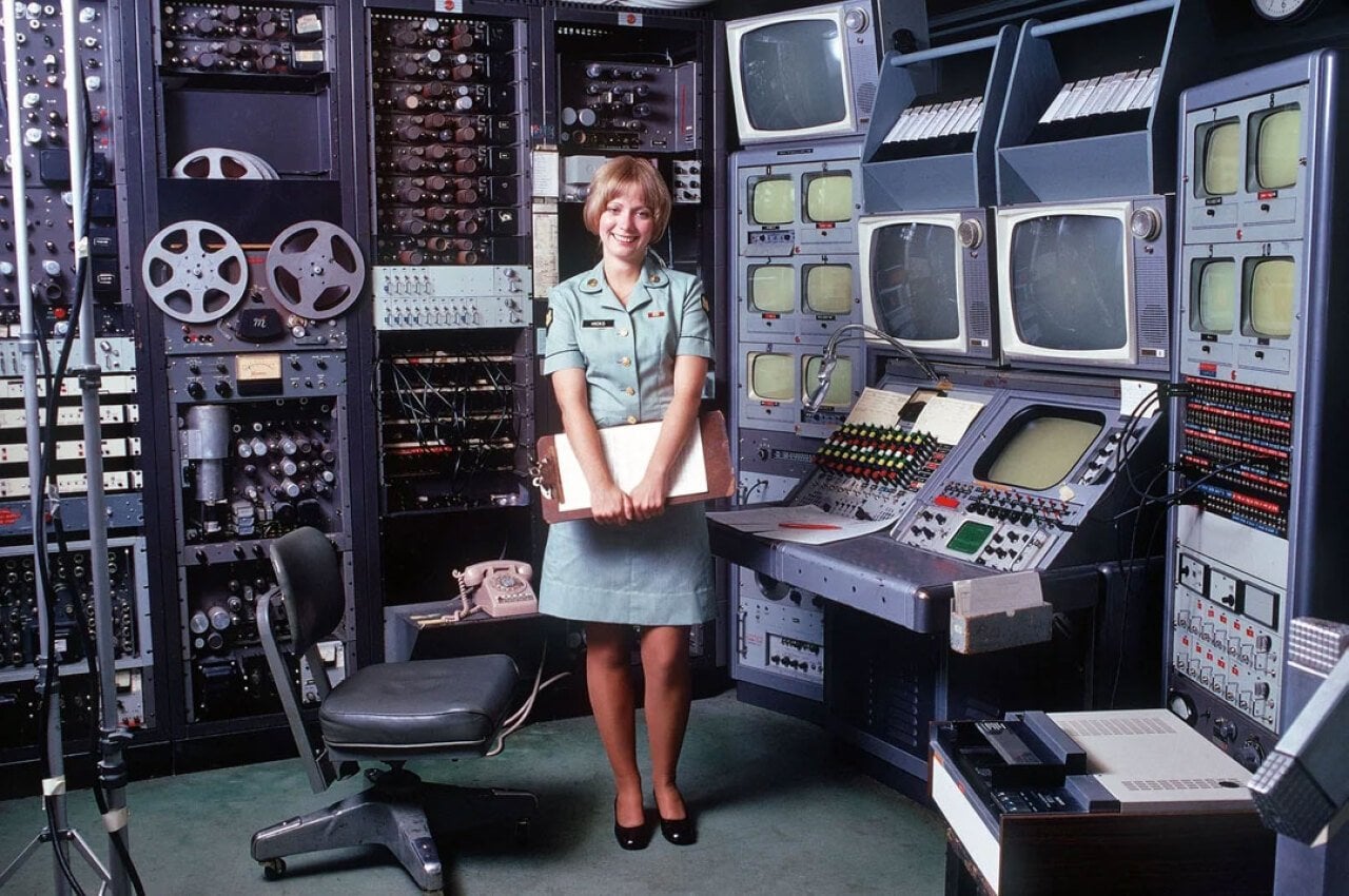 A woman in an Army uniform stands in front of a bays of televisions and audio devices